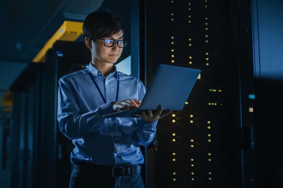 In Dark Data Center Male IT Specialist Stands Beside the Row of Operational Server Racks, Uses Laptop for Maintenance. Concept for Cloud Computing, Artificial Intelligence, Supercomputer, Cybersec